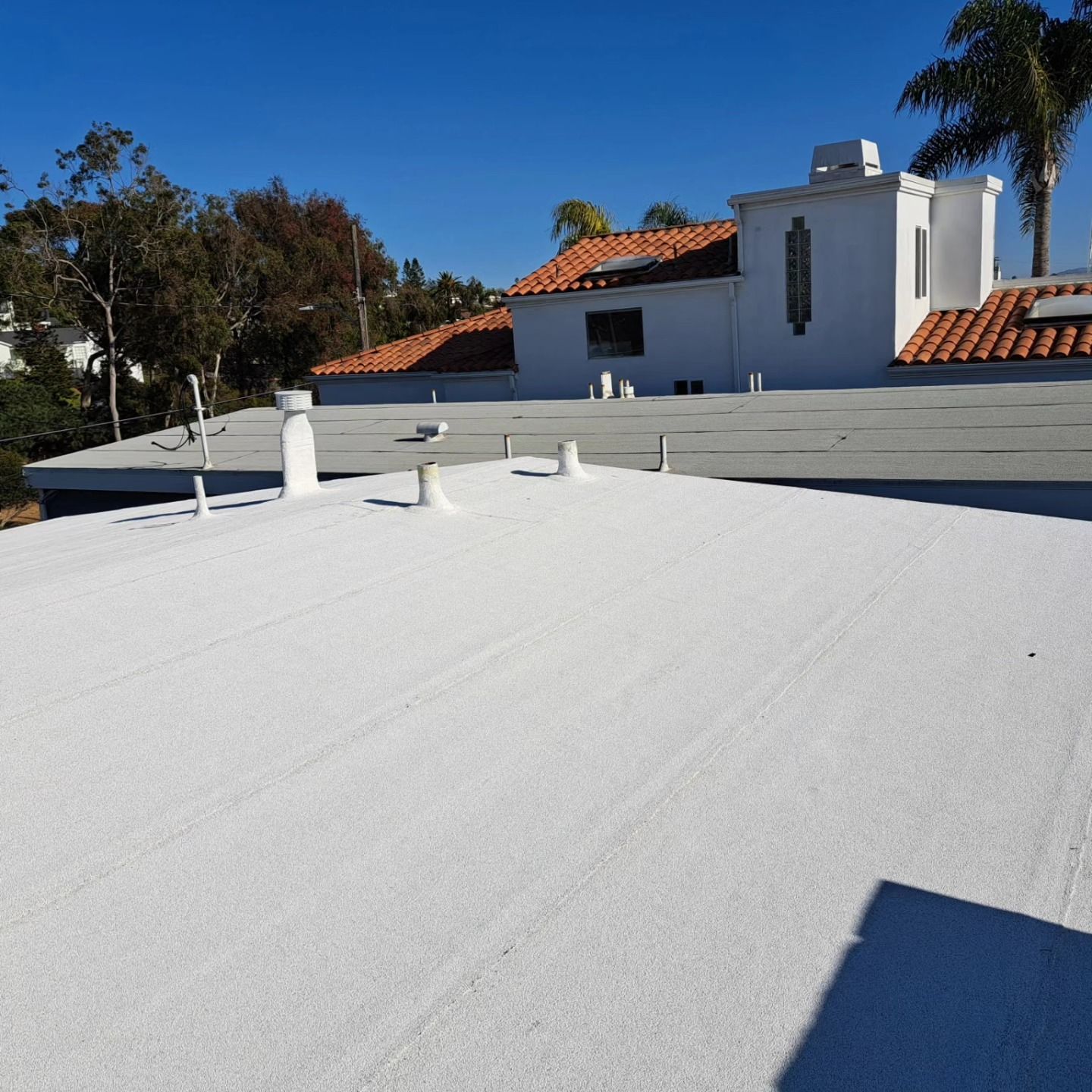 White flat rooftop in foreground with a white stucco house and red tile roofs under a clear blue sky.