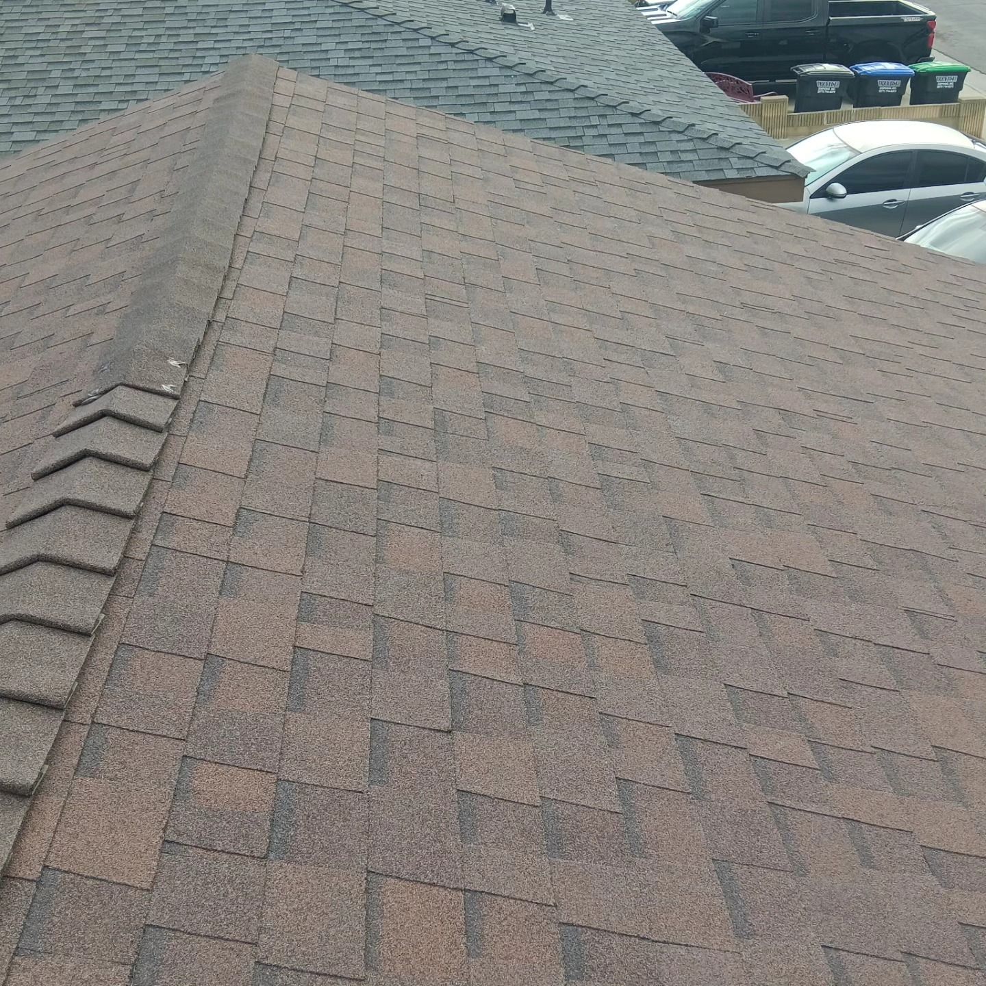 Close-up of a shingled roof with a ridge vent and parked cars visible in the background