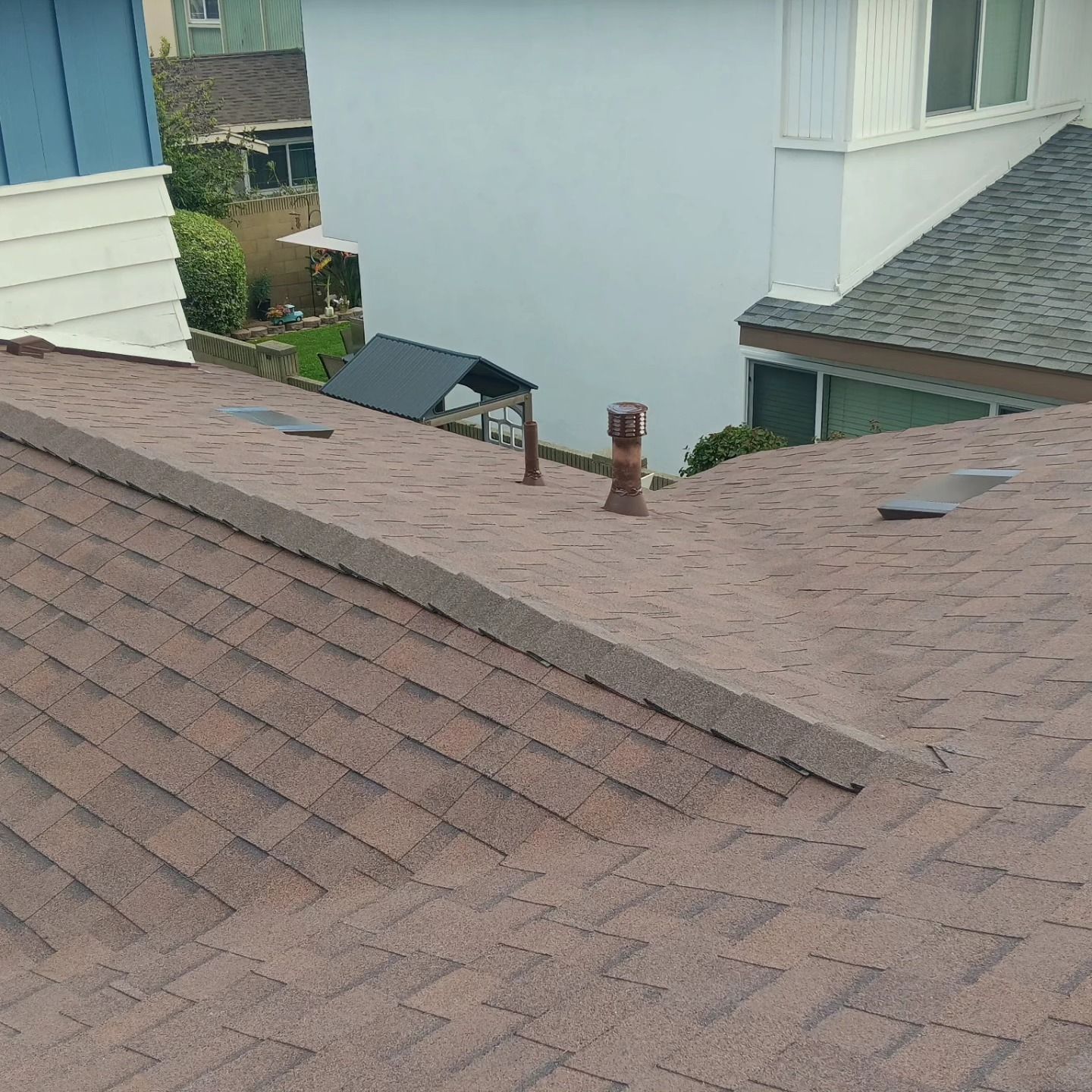 Brown shingled roof with vents and a chimney between neighboring houses