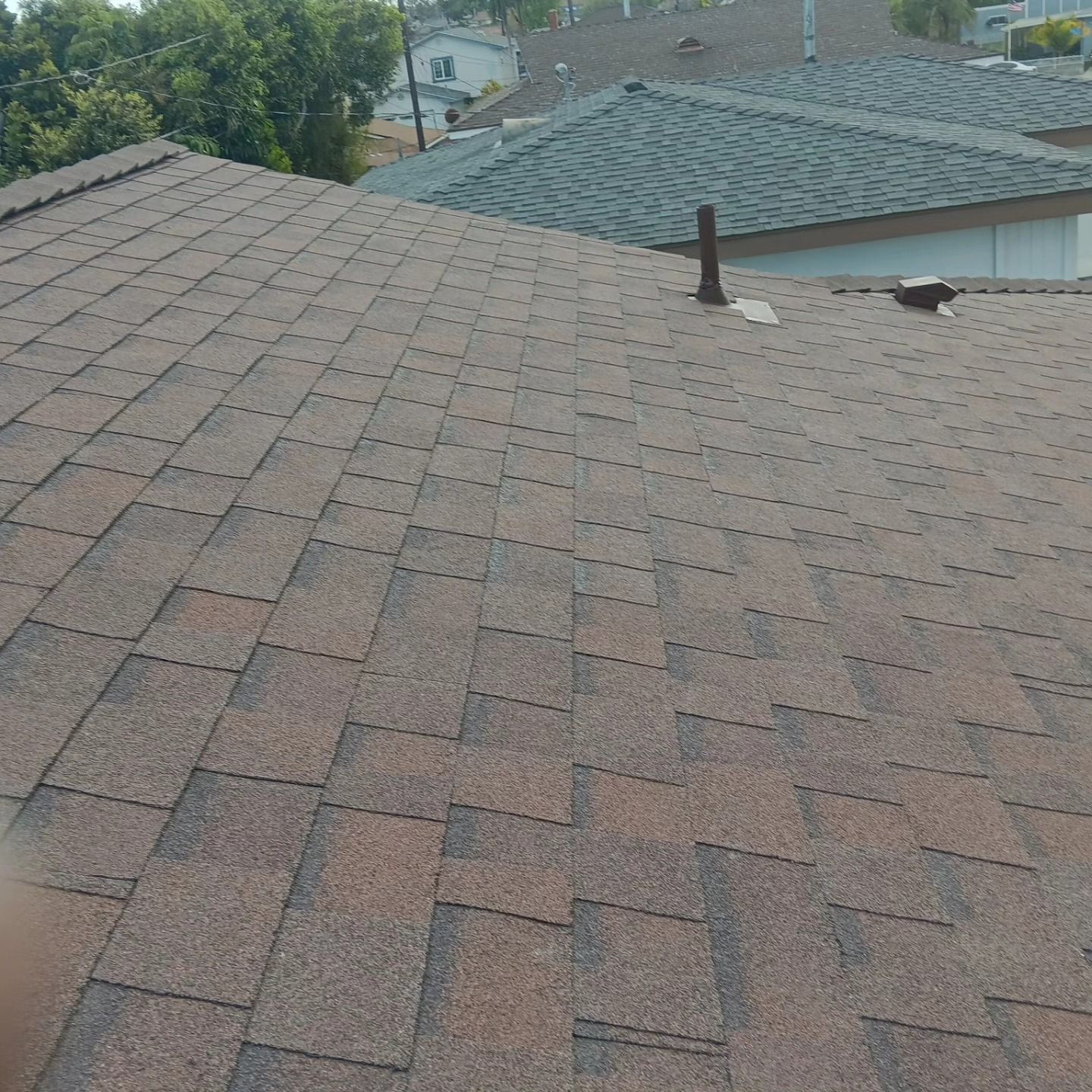 Reddish-brown shingled rooftop with a vent pipe, trees, and neighboring house in the background