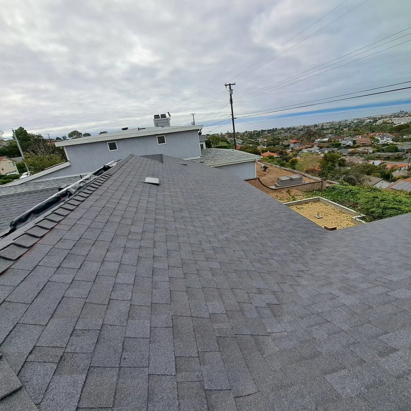 Rooftop with gray shingles, skylight, and distant hillside neighborhood under cloudy sky