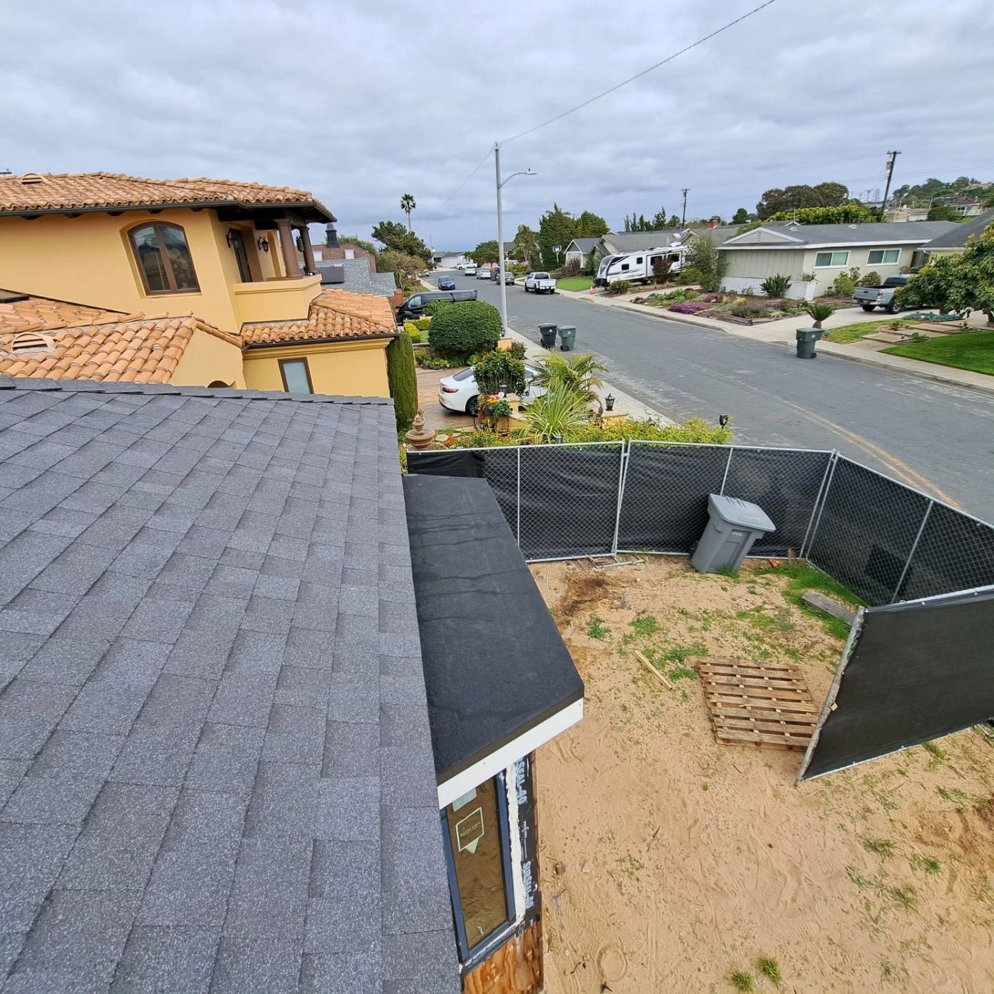 View from a rooftop over a suburban street, with a fenced yard, houses, and cloudy sky.