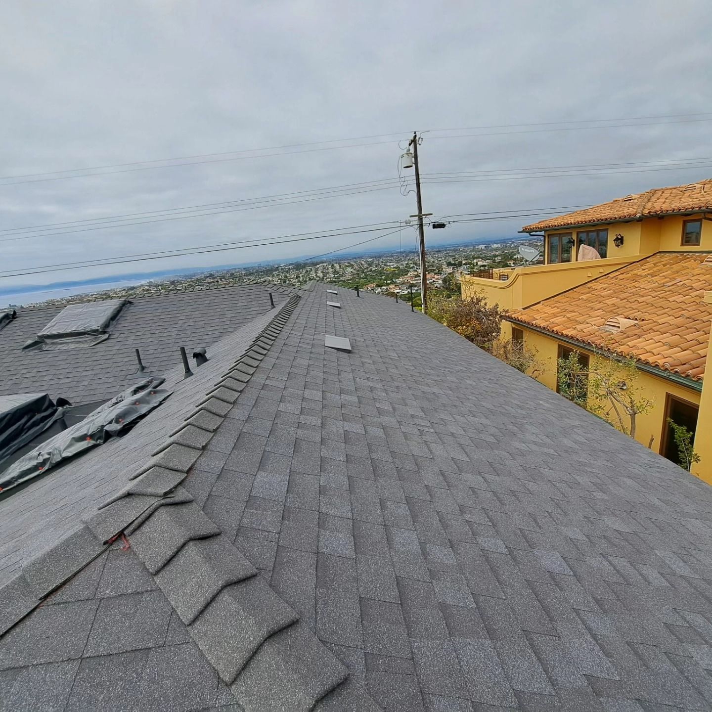 Rooftop with gray shingles beside tan houses, overlooking a cloudy coastal view.