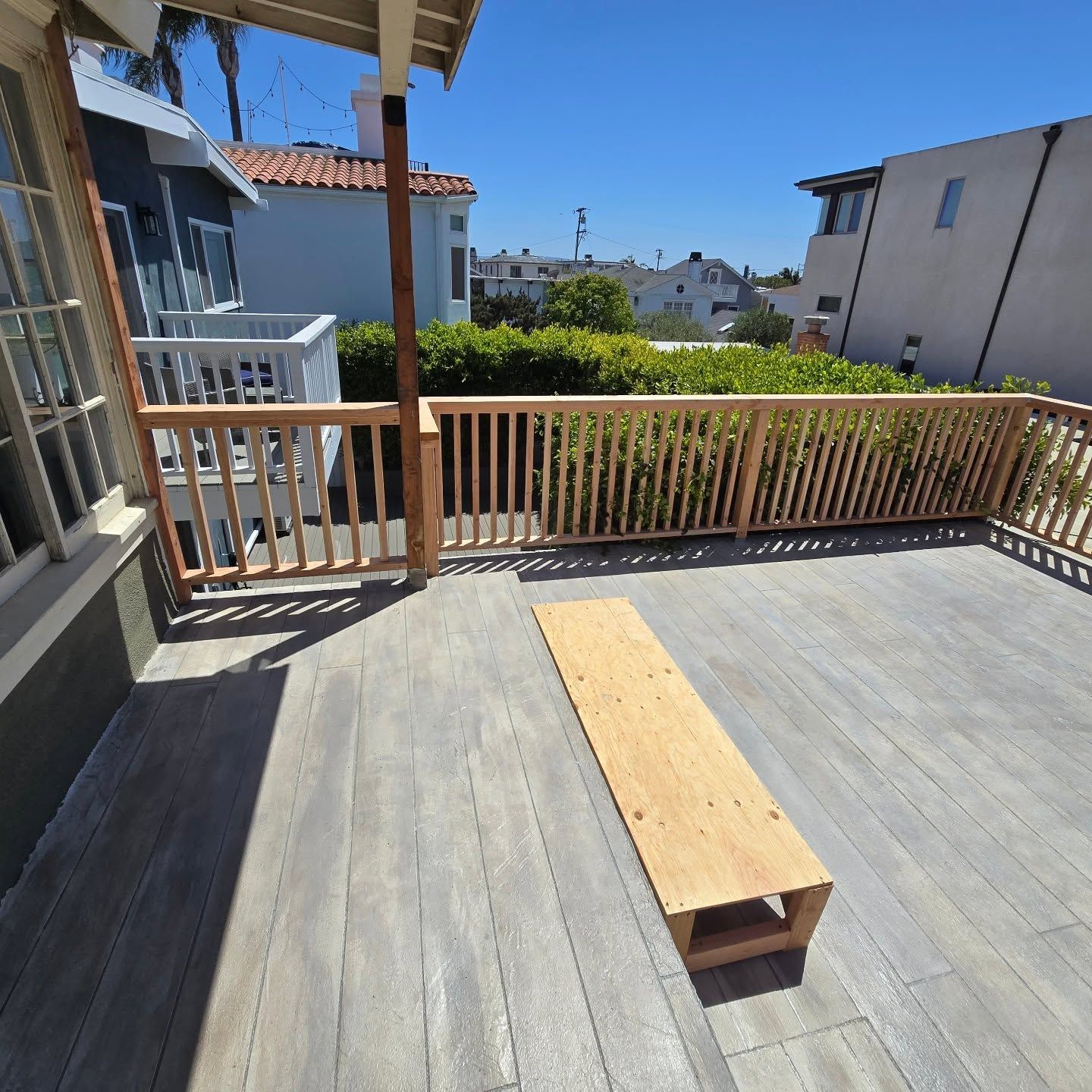 Sunny balcony with gray deck, wooden bench, and railing overlooking neighboring houses and trees.
