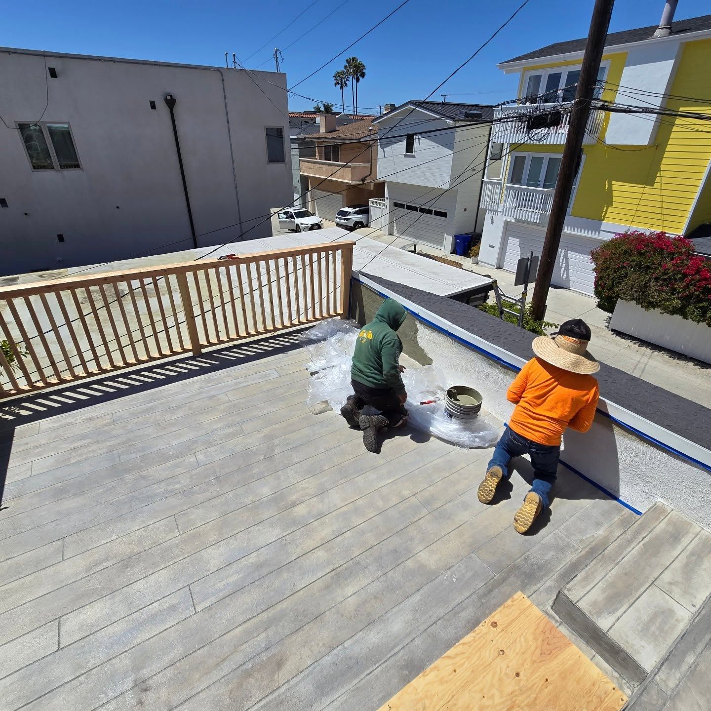 Two workers painting a wooden deck on a sunny rooftop terrace.