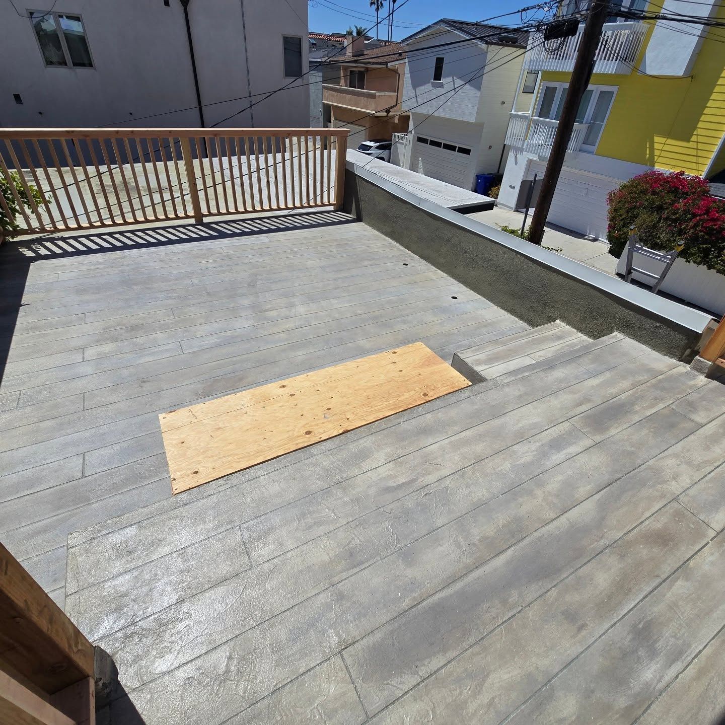 Sunny rooftop deck with gray boards, a wooden ramp panel, and railings overlooking nearby houses