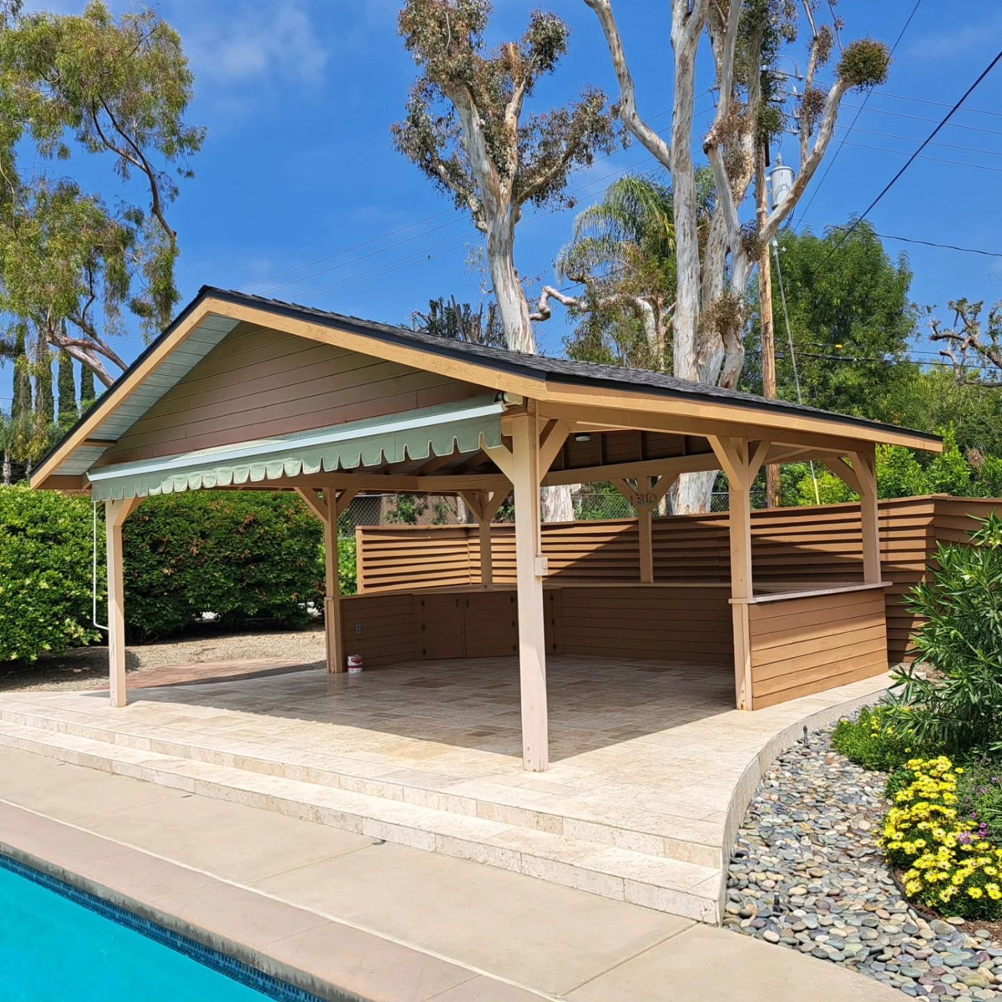 Covered poolside pavilion with wooden posts and sloped roof beside a swimming pool and landscaped yard