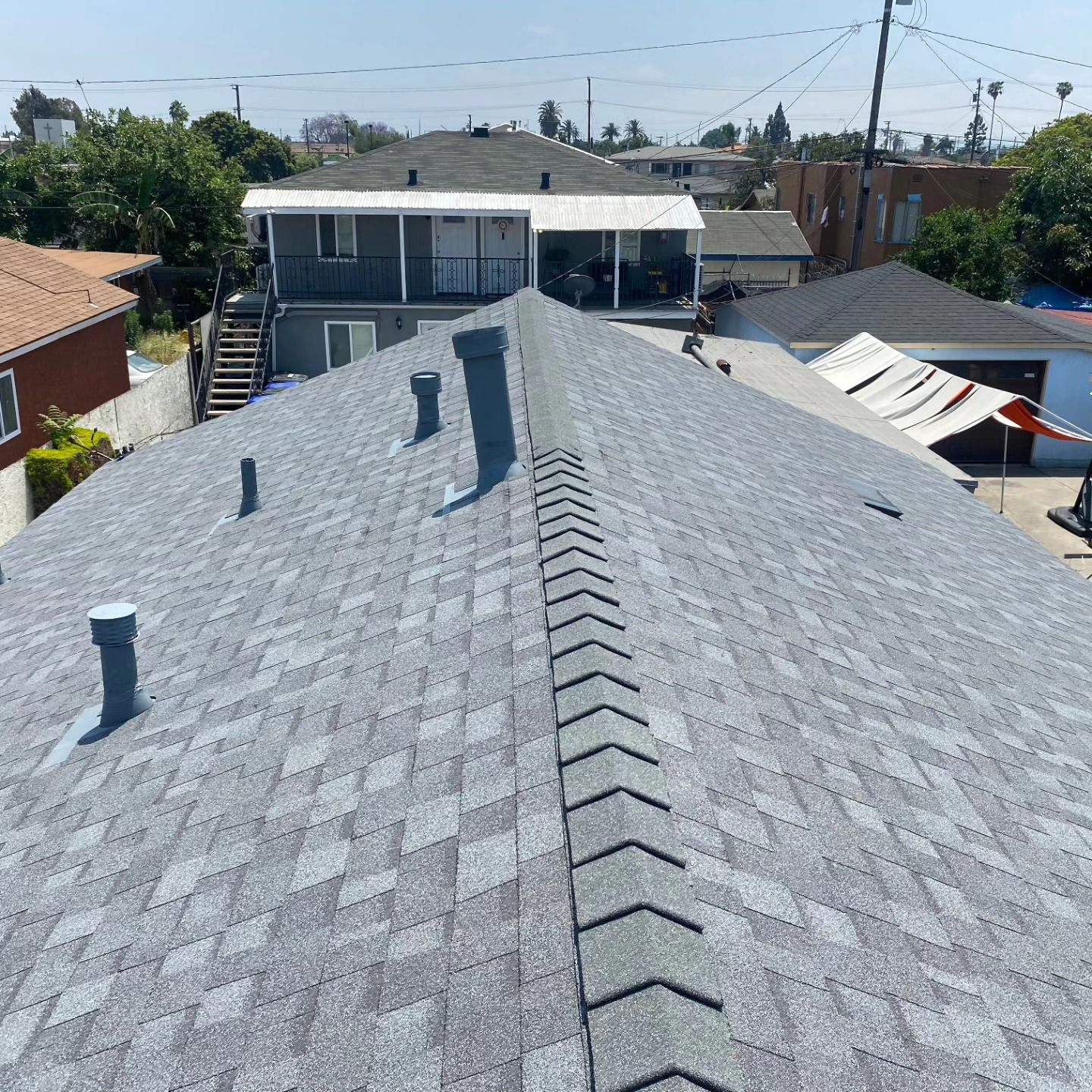 Rooftop view of a gray shingled house with vent pipes, neighboring homes, and a clear blue sky.
