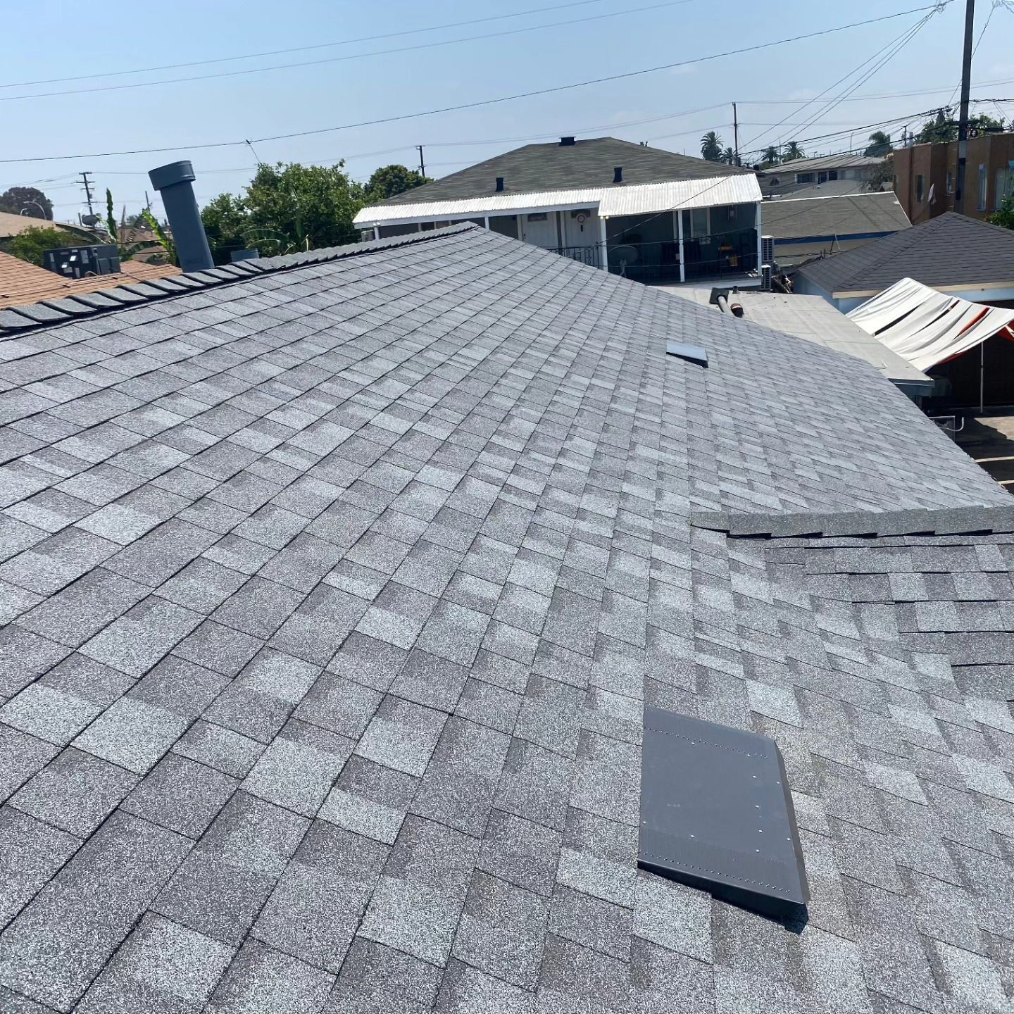 Gray shingled rooftop with a small vent and rooftops, power lines, and sky in the background
