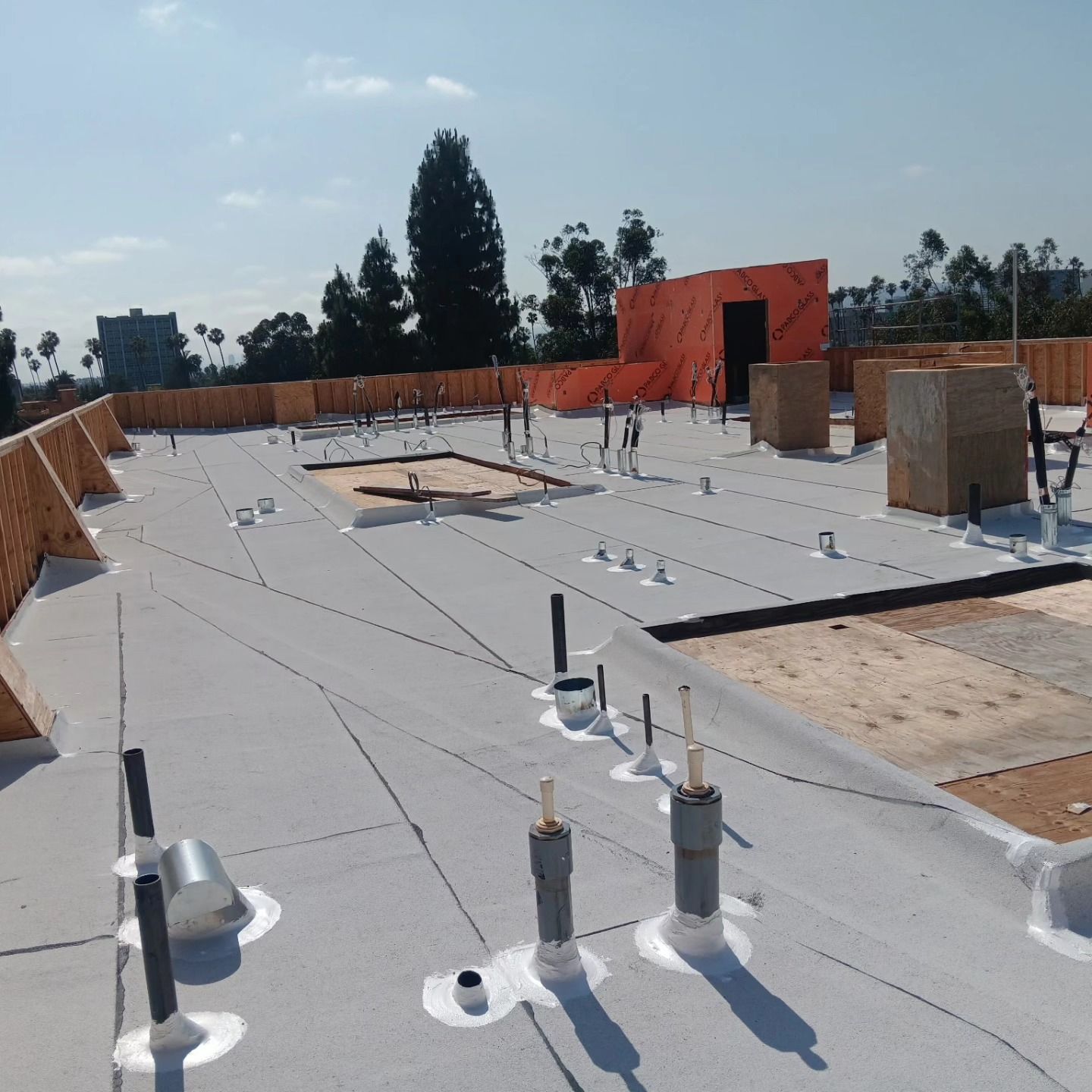 Flat rooftop under construction with white membrane, pipes, vents, and orange parapet walls.