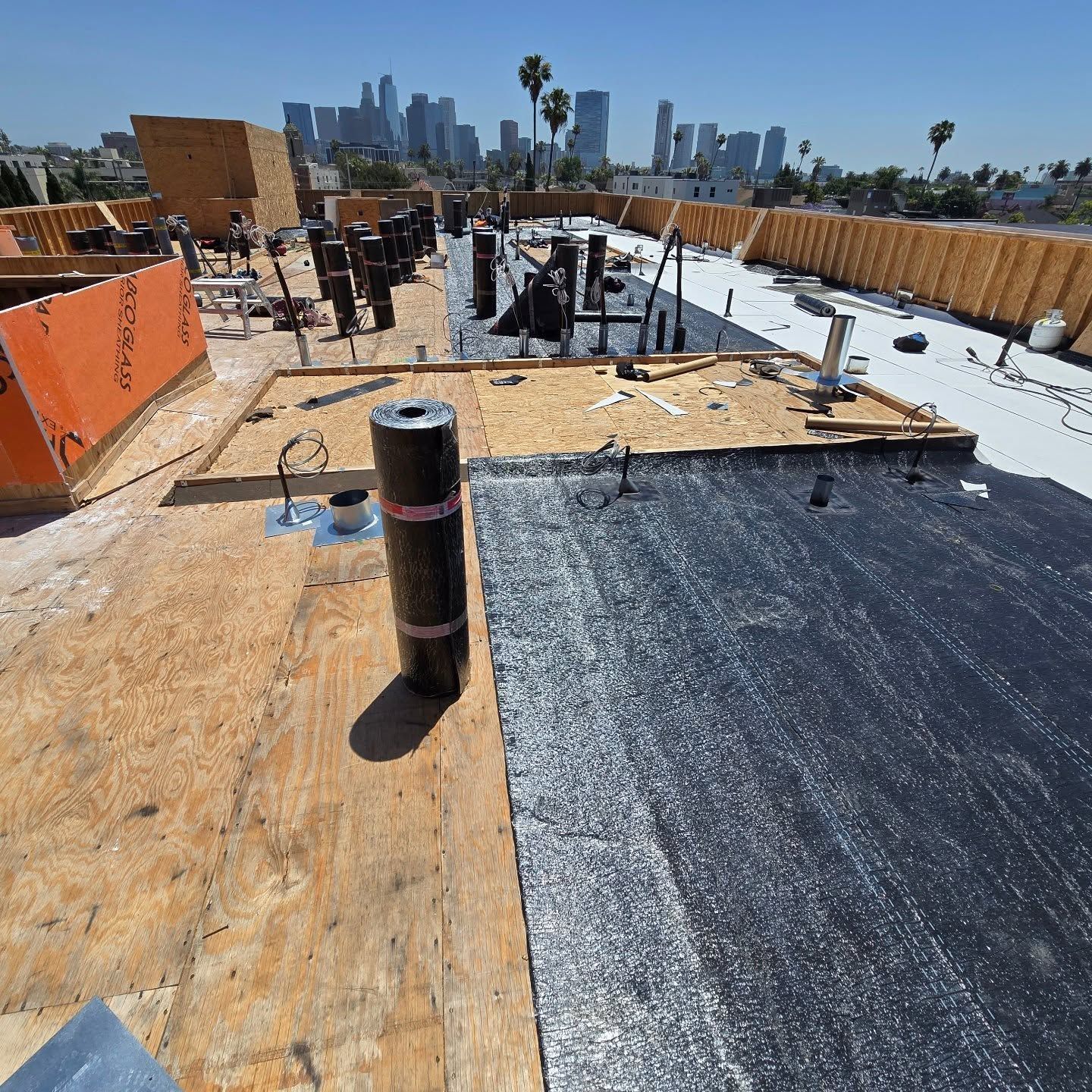 Rooftop under construction with plywood, black roofing membrane, and city skyline in the background