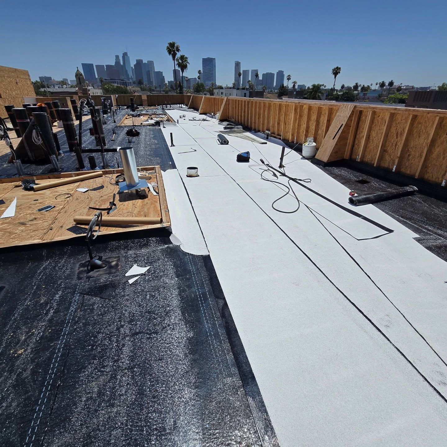 Rooftop under construction with workers, white roofing membrane, and a city skyline in the distance