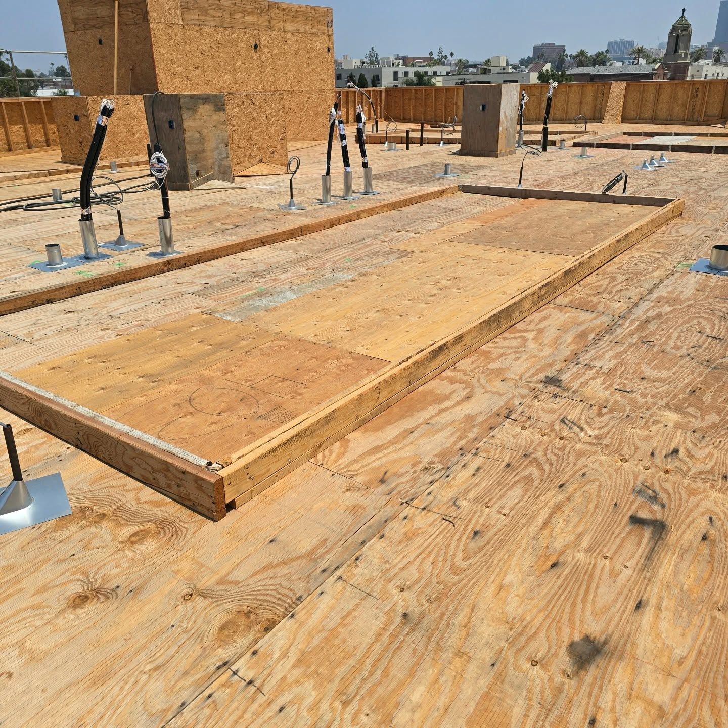 Wooden rooftop construction deck with workers, plumbing pipes, and low brick structures under a clear sky