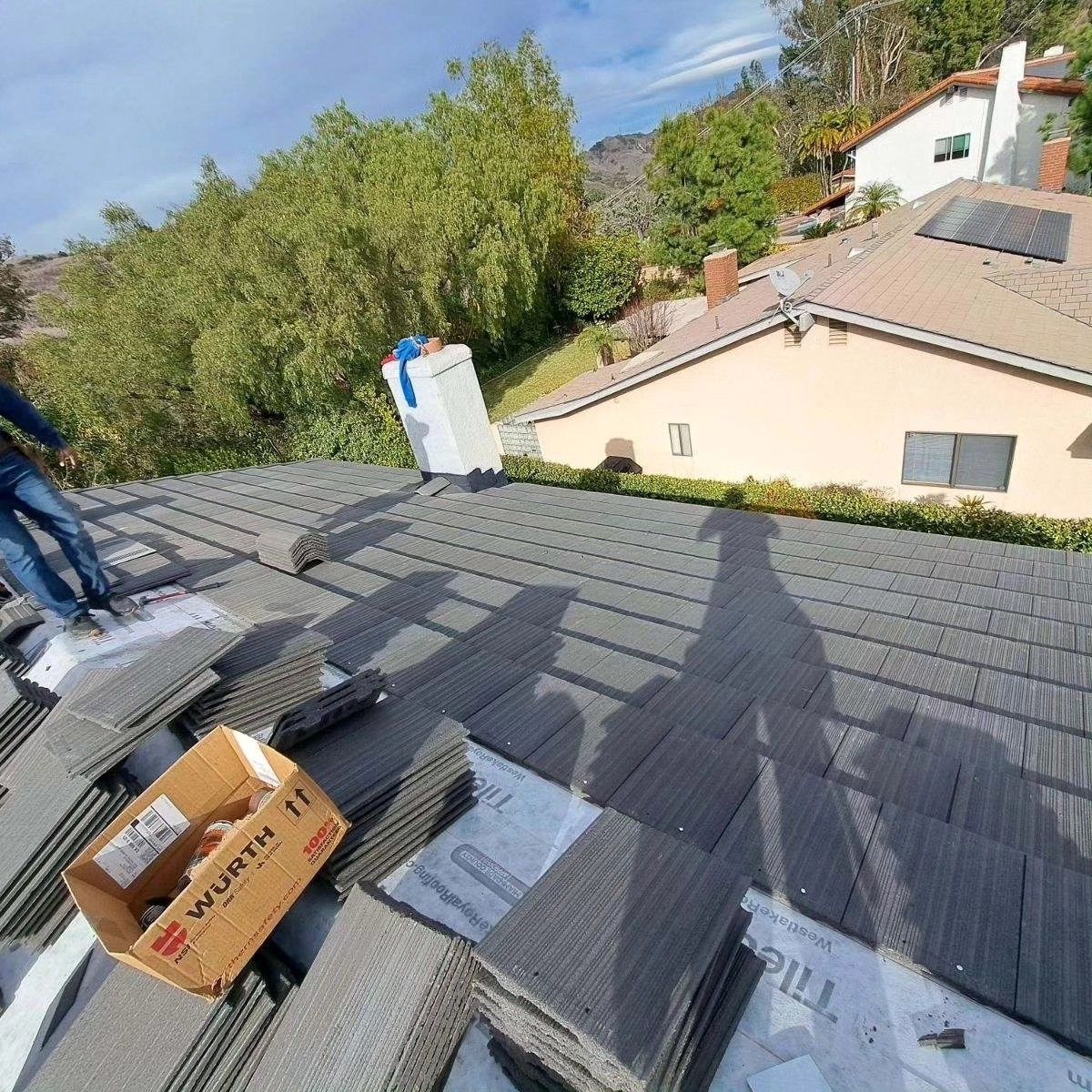 Rooftop with dark shingles, boxes, and roofing materials beside a house and trees on a sunny day
