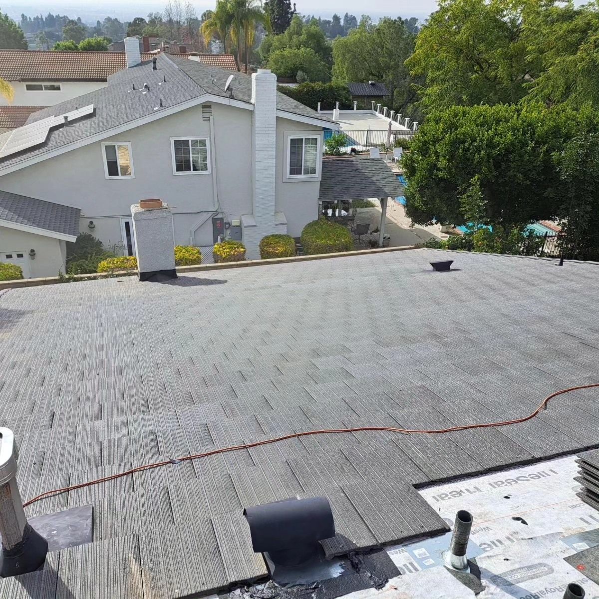 Roof view with gray shingles, nearby houses, trees, and a blue sky in the background