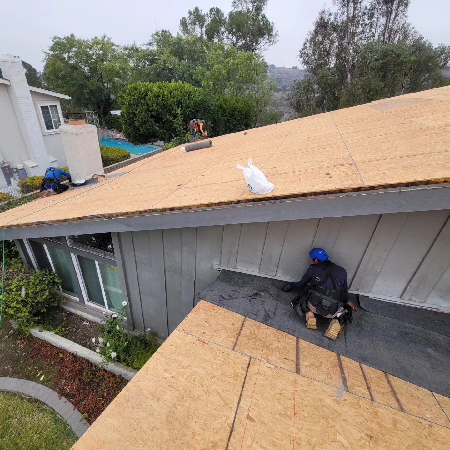 Workers installing a tan roof on a gray house, with one worker on the roof and one below.