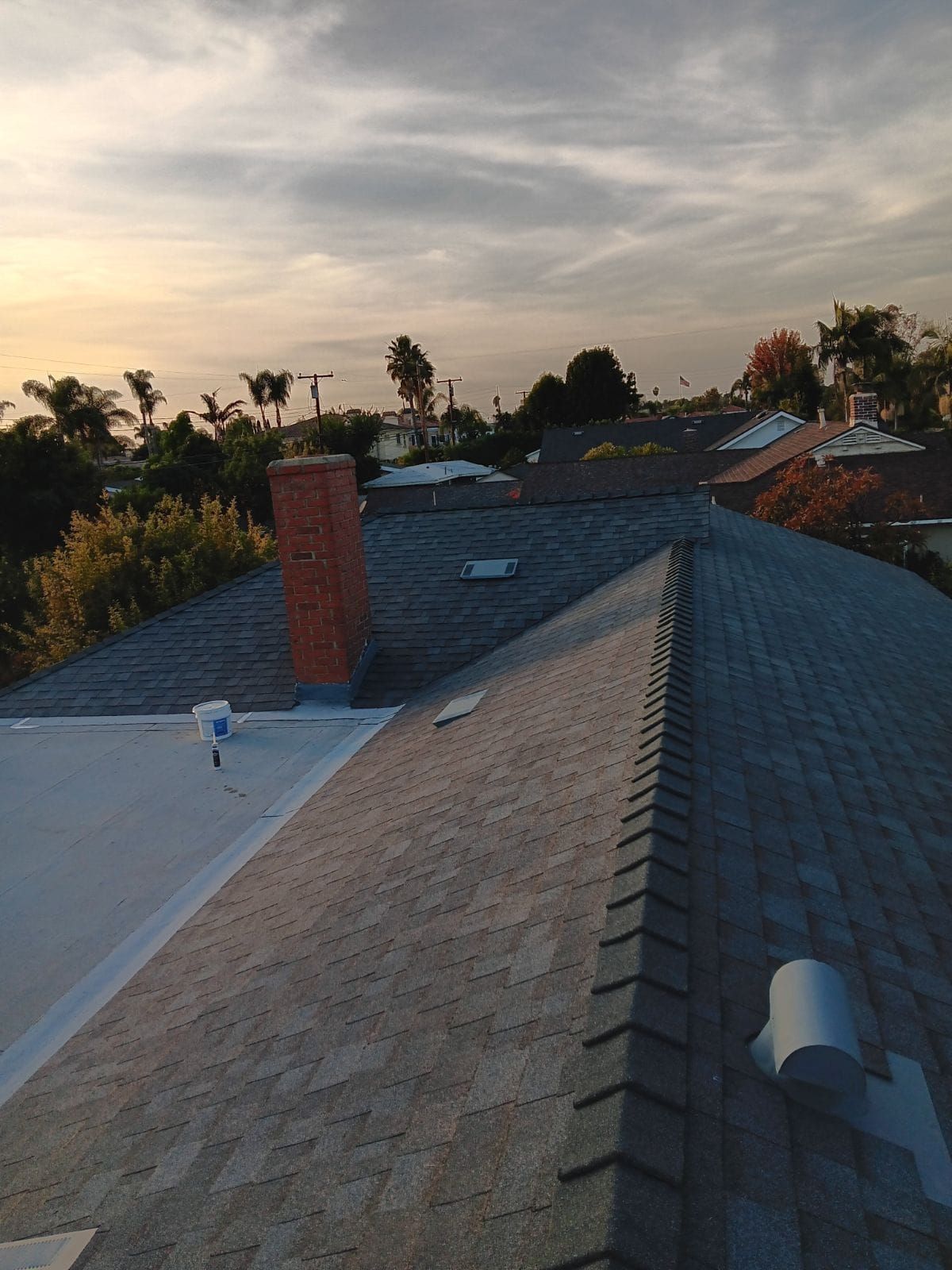 Roofscape at sunset with gray shingles, white flat roof, brick chimney, and trees in the distance
