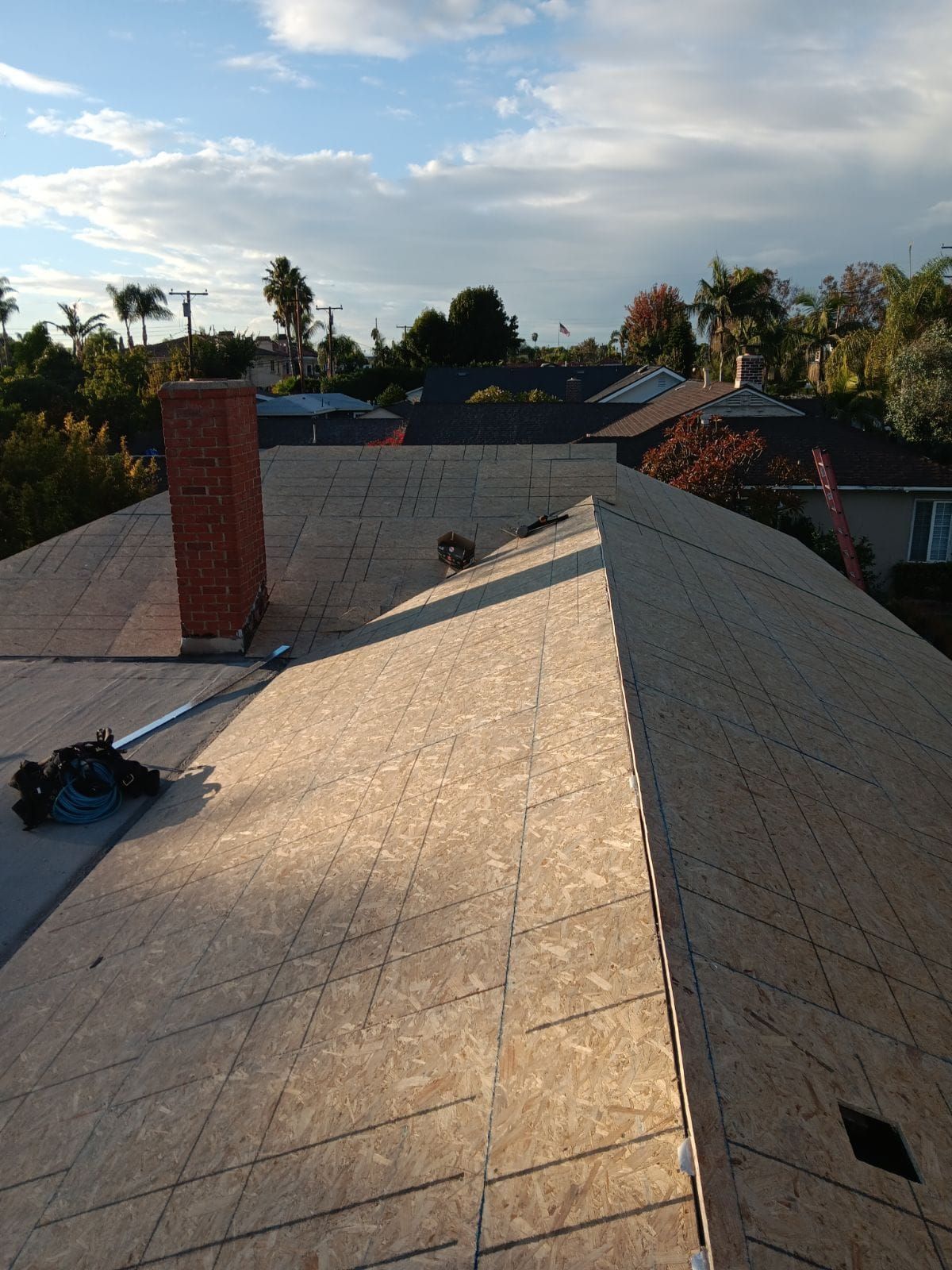 Sunlit rooftop under construction with shingles, chimney, and trees in the distance.