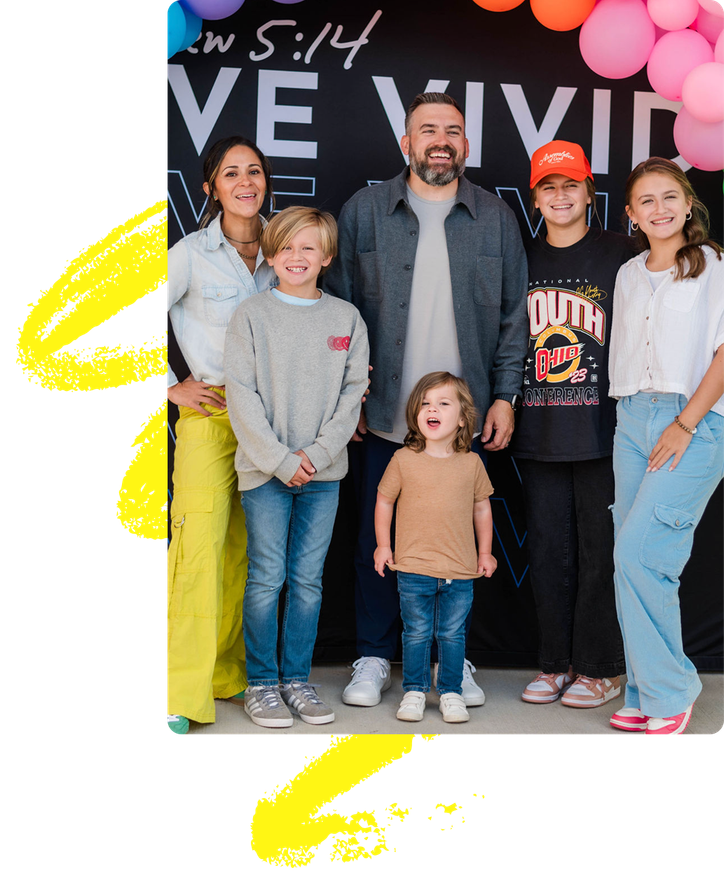 Family of six posing in front of a pink balloon backdrop with “WE WILL” text