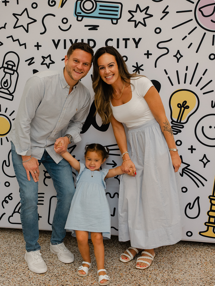 Family posing smiling in front of a doodle mural, with a child between two adults holding hands.