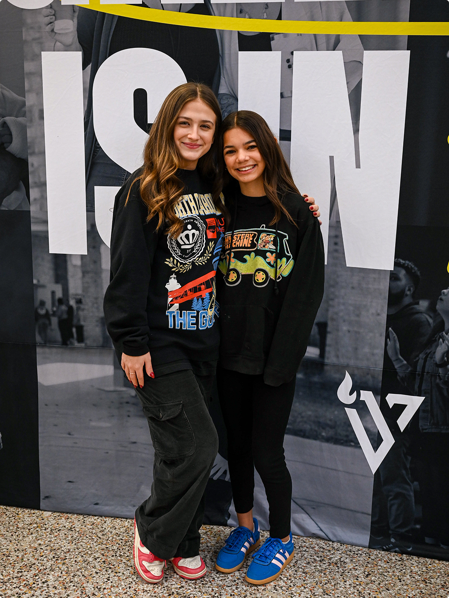 Two people smiling and posing in front of a large “SHOW” backdrop, wearing black graphic tops and sneakers.