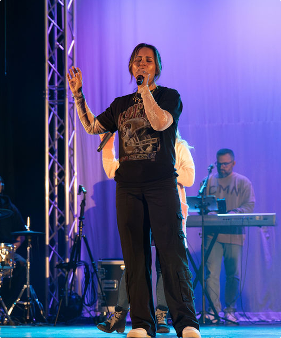 Singer on stage holding a microphone, with a keyboardist in the background under purple lighting