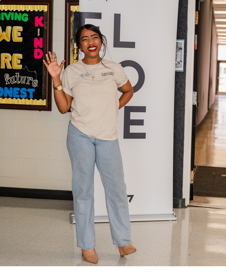 Woman smiling and waving in a hallway, wearing a beige shirt and light blue jeans.