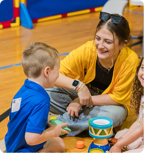 Children playing with a smiling adult on a gym floor, sharing a drum and toys.