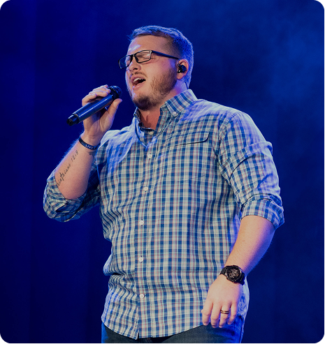 Singer in a blue plaid shirt performing on stage with a microphone under blue lighting