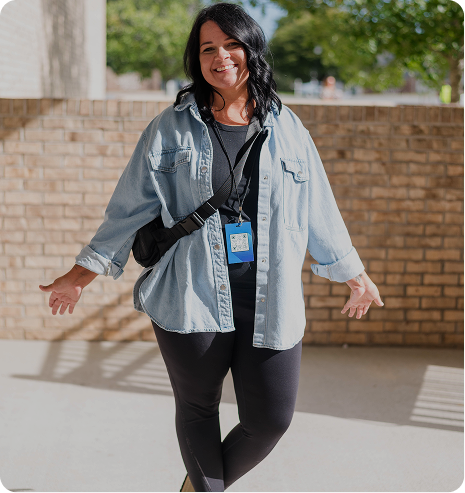 Smiling person in a light denim shirt and black leggings standing outdoors in front of a brick wall.