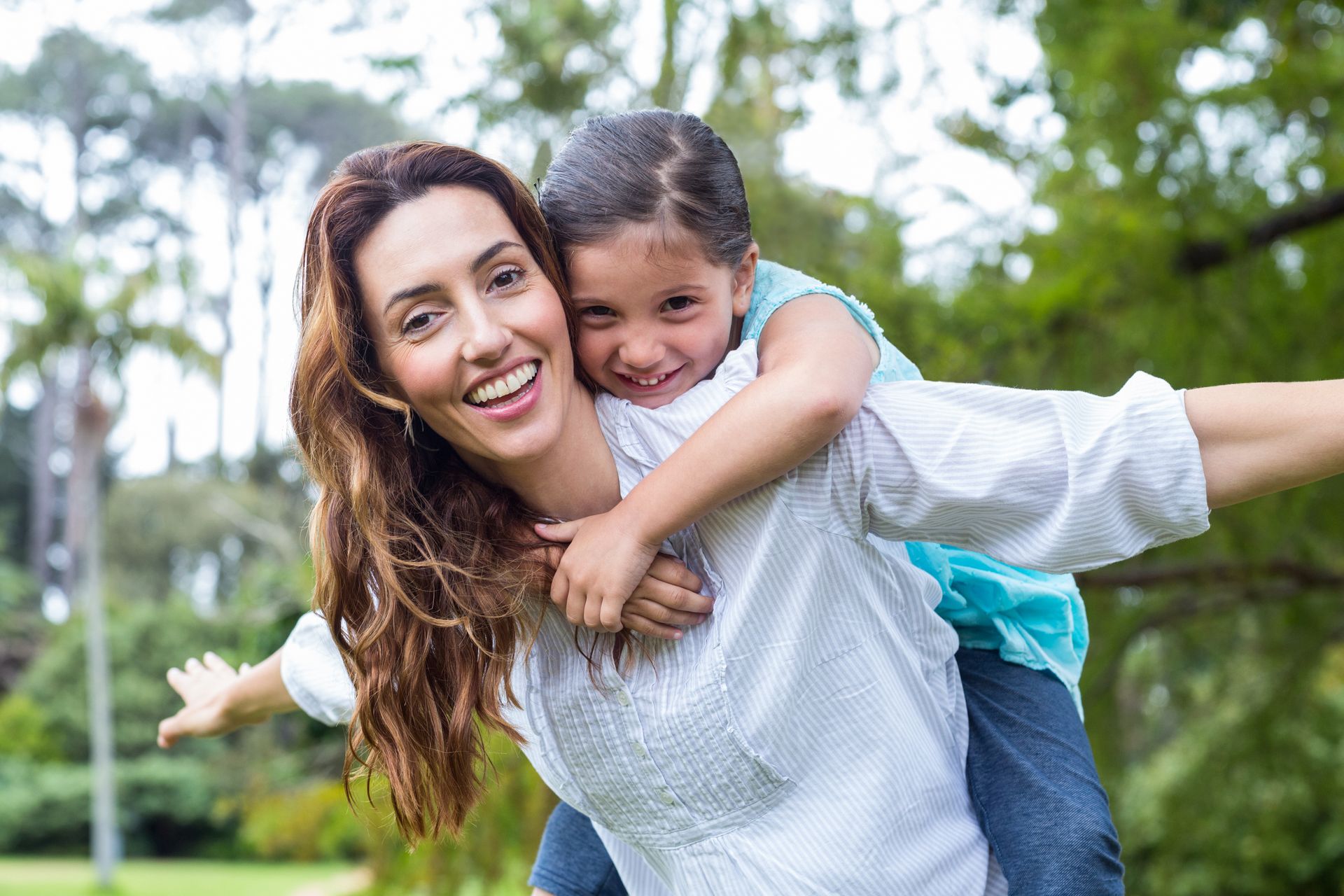 A woman is in a park and has a child on her back. They are both smiling.