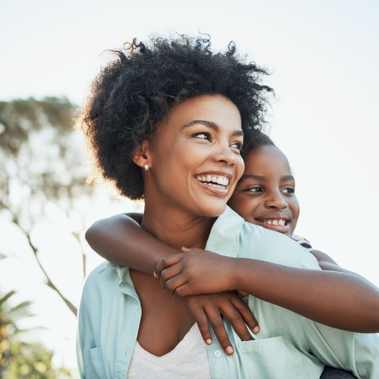 A child is hugging their mother from behind, with a tree in the background. They are both smiling.