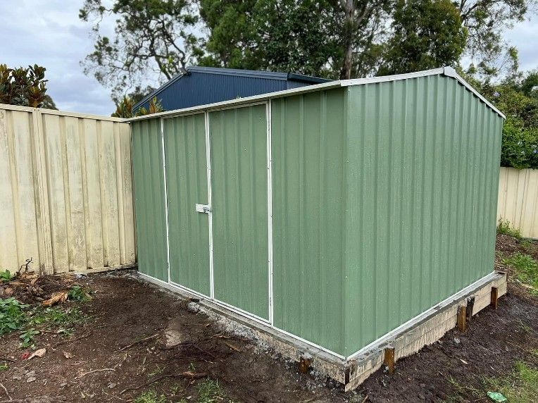 A gReen Metal Shed is Sitting in the Dirt Next to a Fence — JOA - Construction & Maintenance Pty Ltd in Appin, NSW
