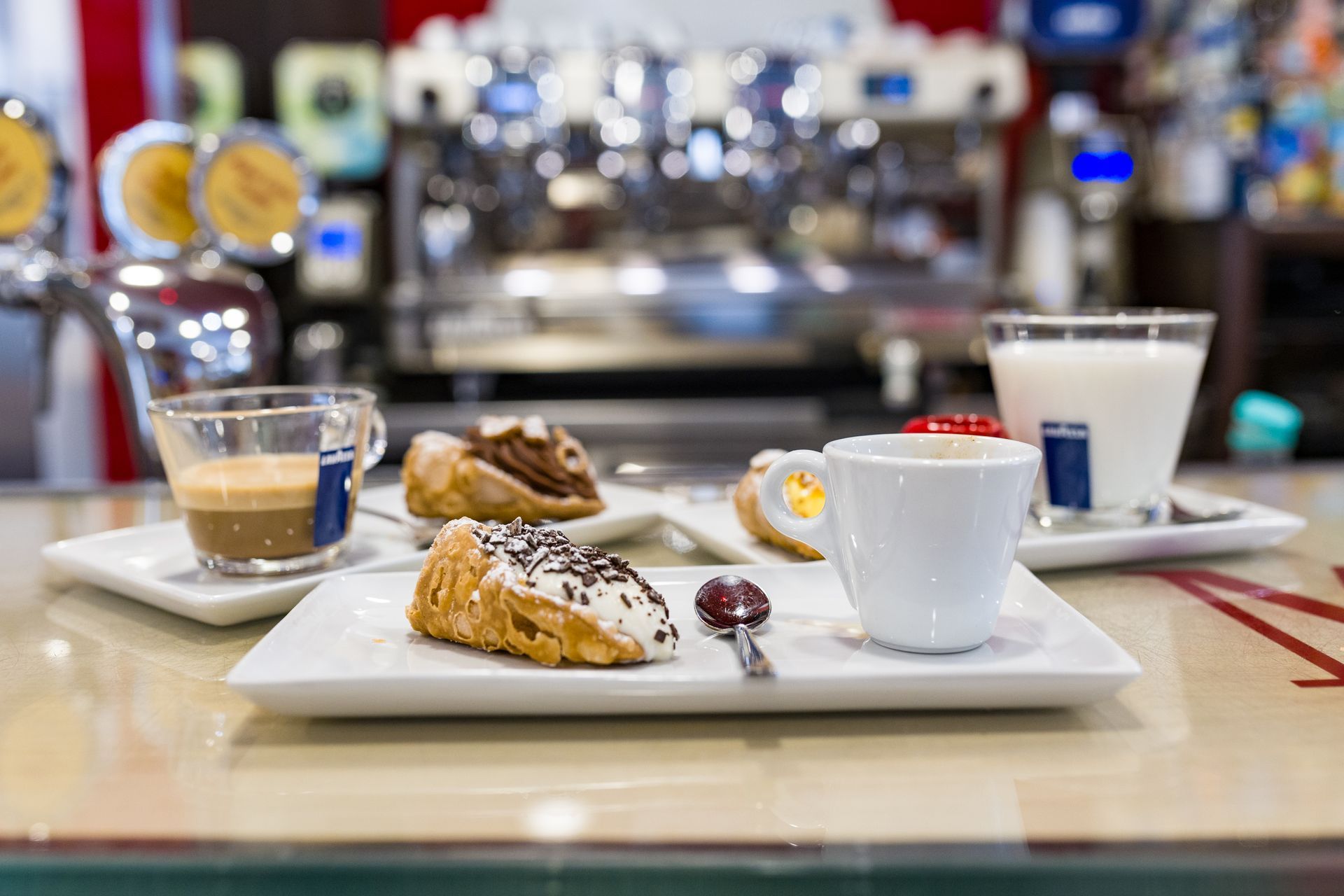 Caffè e pasticcini sul bancone di un bar, con una tazza bianca, un espresso e un bicchiere di latte sullo sfondo.