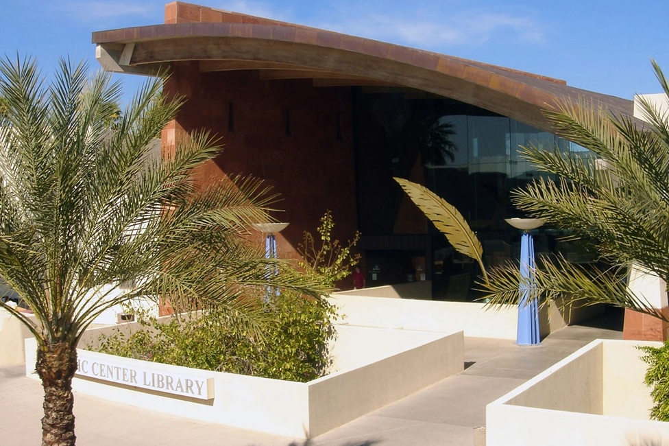 Modern library entrance with curved roof, glass facade, white walls, and palm trees in front