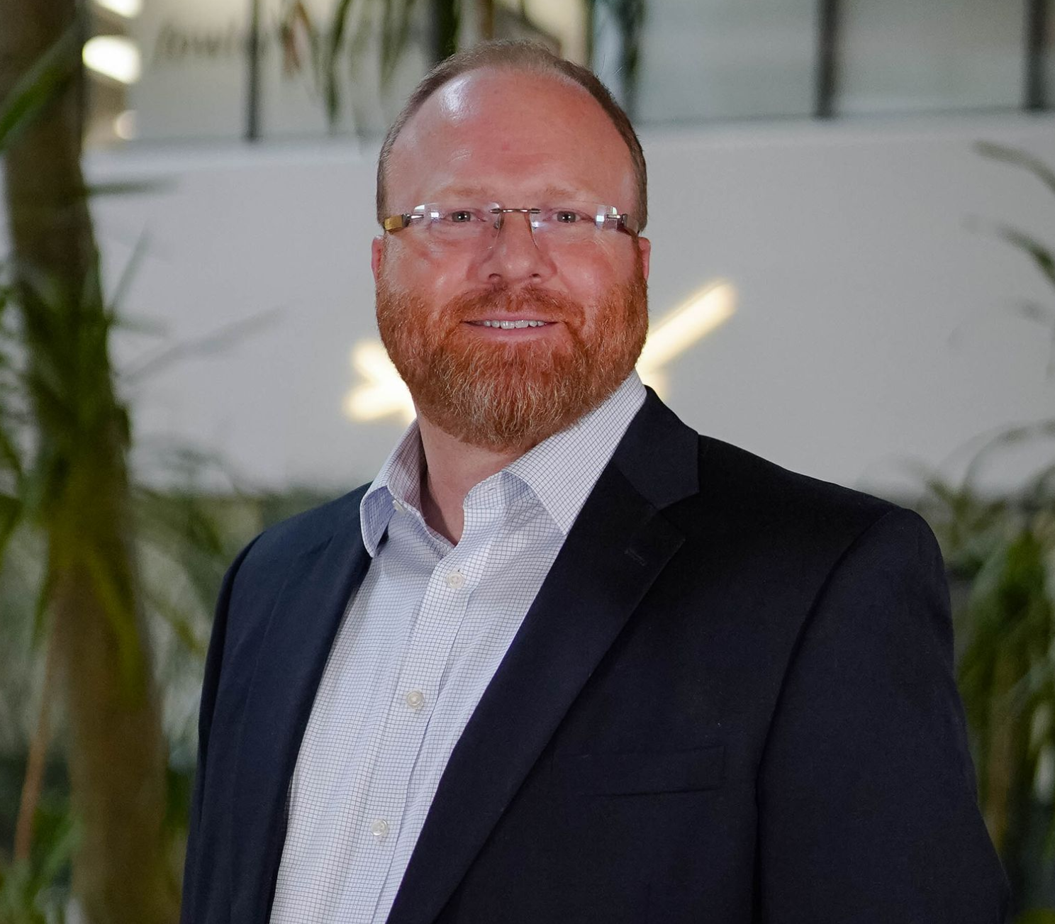 Smiling man in glasses and a dark suit jacket standing indoors with plants in the background