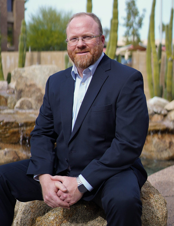 Man in navy suit seated outdoors on a rock, with a waterfall and desert plants behind him.