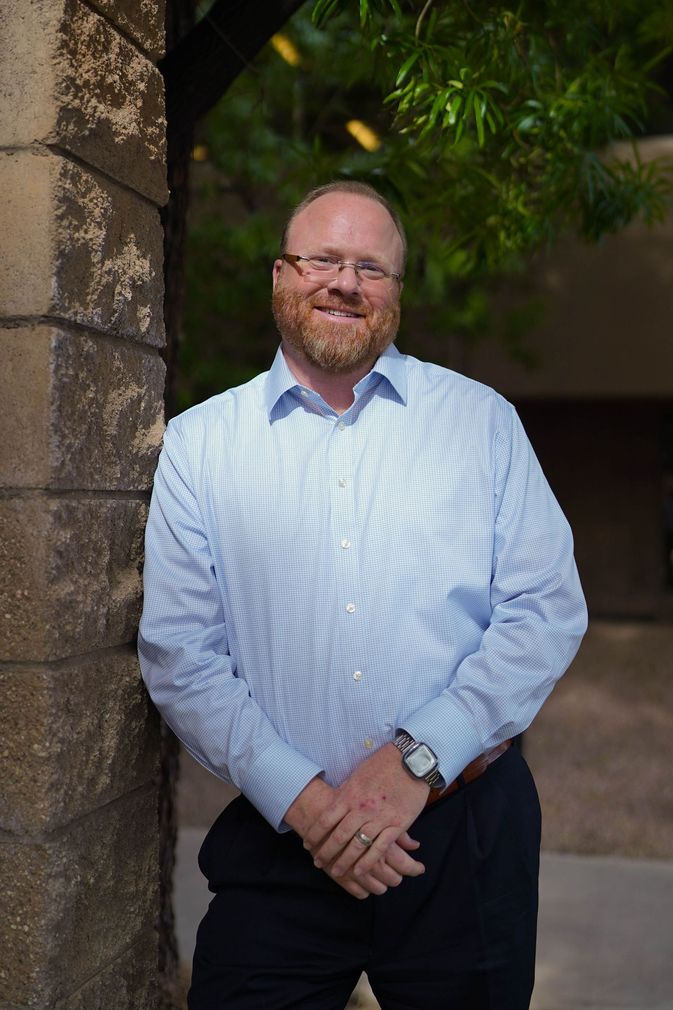 Man standing outdoors by a stone wall, wearing a light blue patterned shirt and dark pants, smiling.