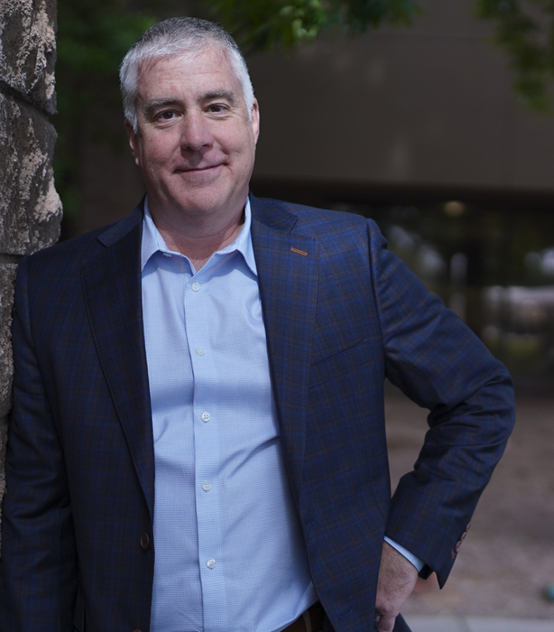 Man in a navy blazer and light blue shirt stands outdoors with one hand on his hip, smiling.