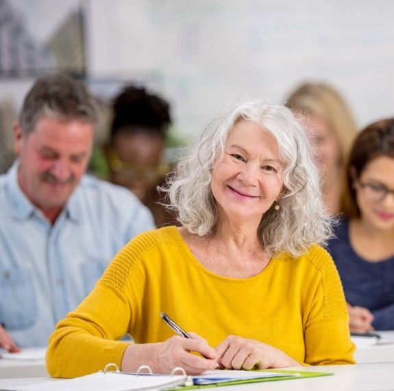Smiling woman in a yellow sweater writing at a desk in a classroom setting