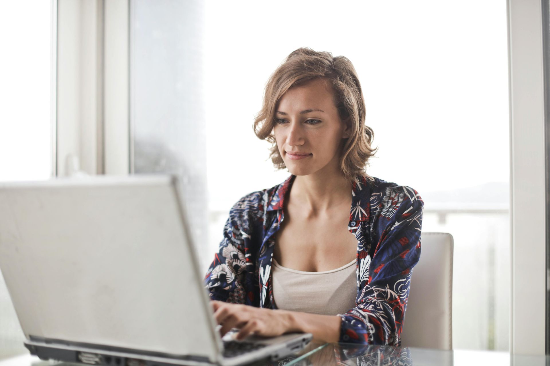Woman typing on a laptop at a table by a bright window