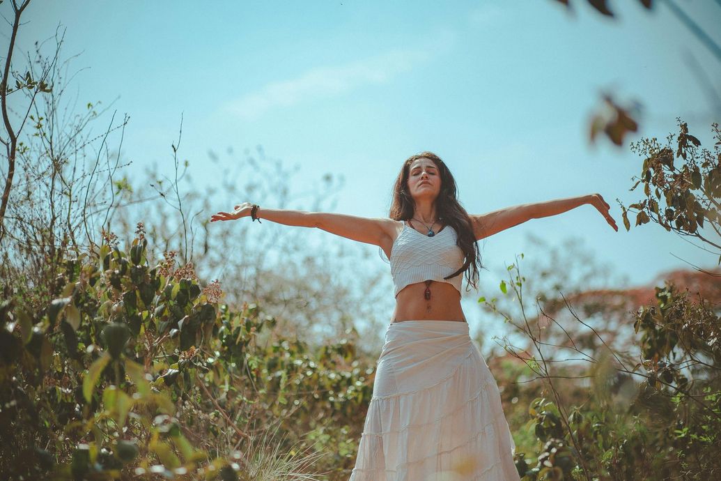 A woman in a white dress is standing in a field with her arms outstretched.
