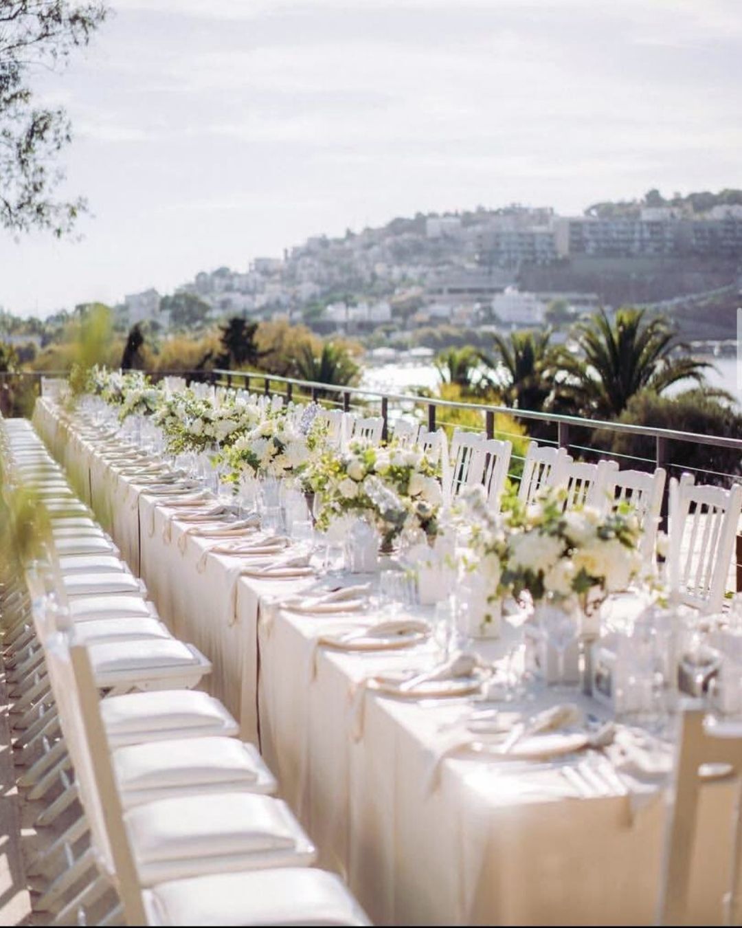 Elemanta Boutique Hotel | Long banquet table set with white chairs and floral arrangements overlooking a waterfront hillside.