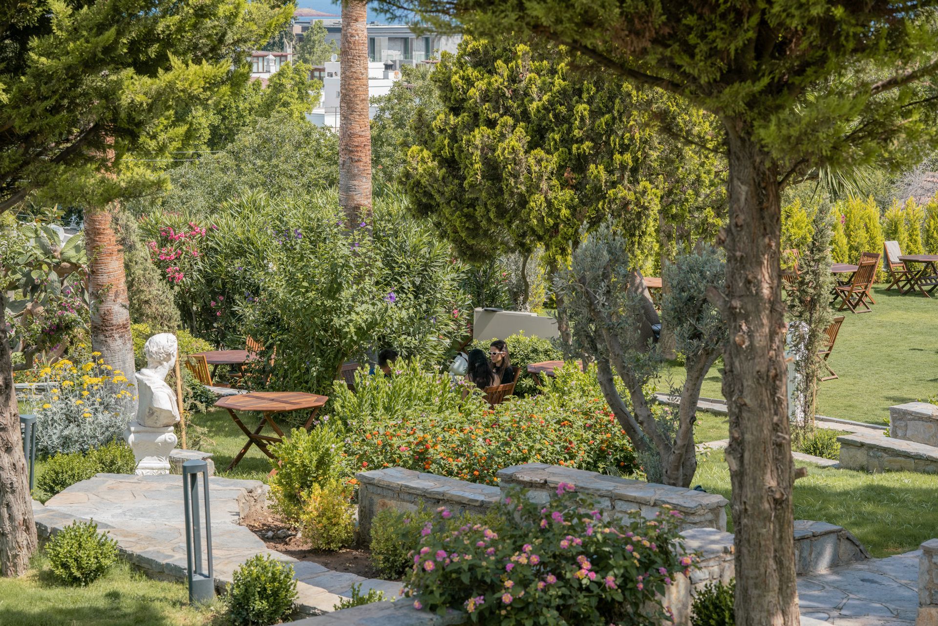 Elemanta Boutique Hotel | Garden path with white statue, stone benches, and flowering greenery under tall trees