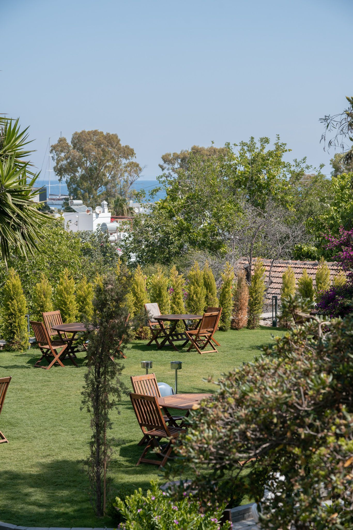 Elemanta Boutique Hotel | Lush garden patio with wooden tables and chairs on a green lawn, bordered by shrubs and trees