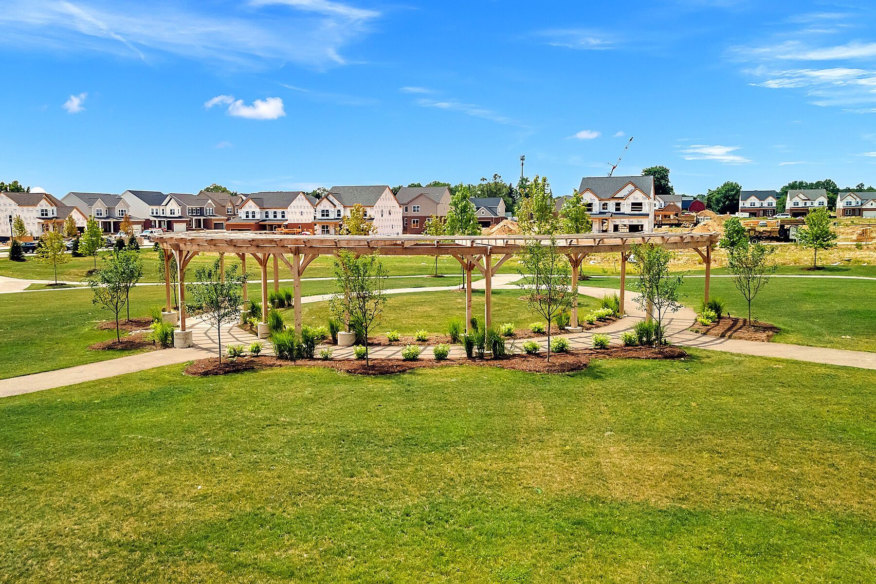 Sunny suburban park with a pergola, flower beds, green lawn, and houses in the background