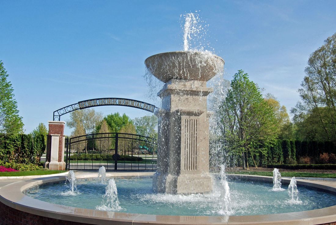 Decorative fountain in a park with water jets, trees, and a pedestrian bridge under a blue sky
