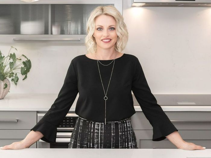 Woman standing in a bright kitchen, smiling with hands on the counter, wearing a black top and patterned skirt
