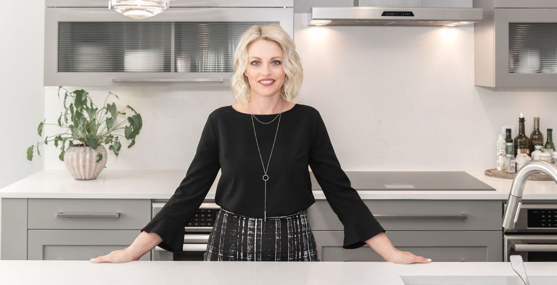 Woman in a modern kitchen, smiling behind a white counter, with gray cabinets and a potted plant.