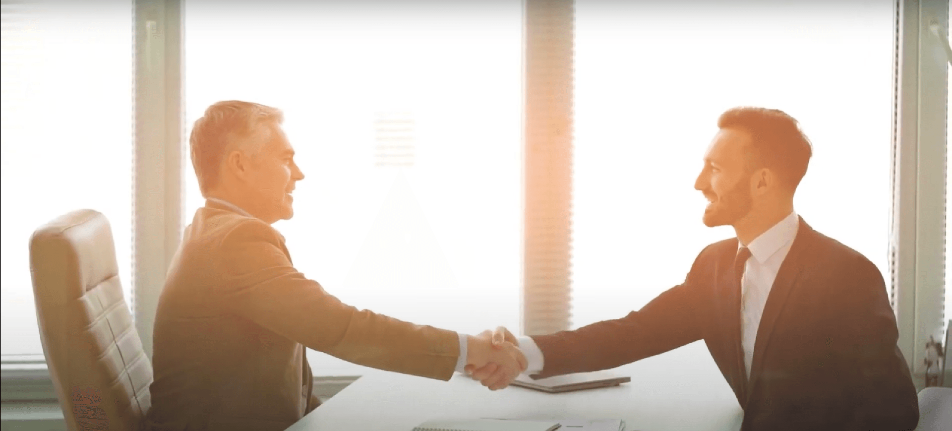Two businesspeople in suits shaking hands across a desk in a bright office
